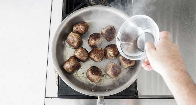 adding water to a pan of meatballs